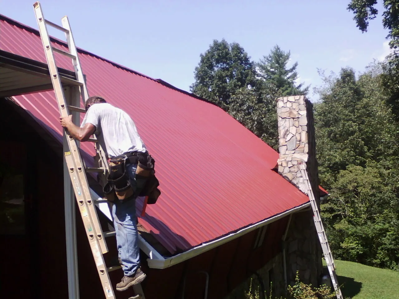 Roofer on a ladder beside a red metal roof and stone chimney — Soffit & Fascia Repair by Roofing Brothers in Charlotte